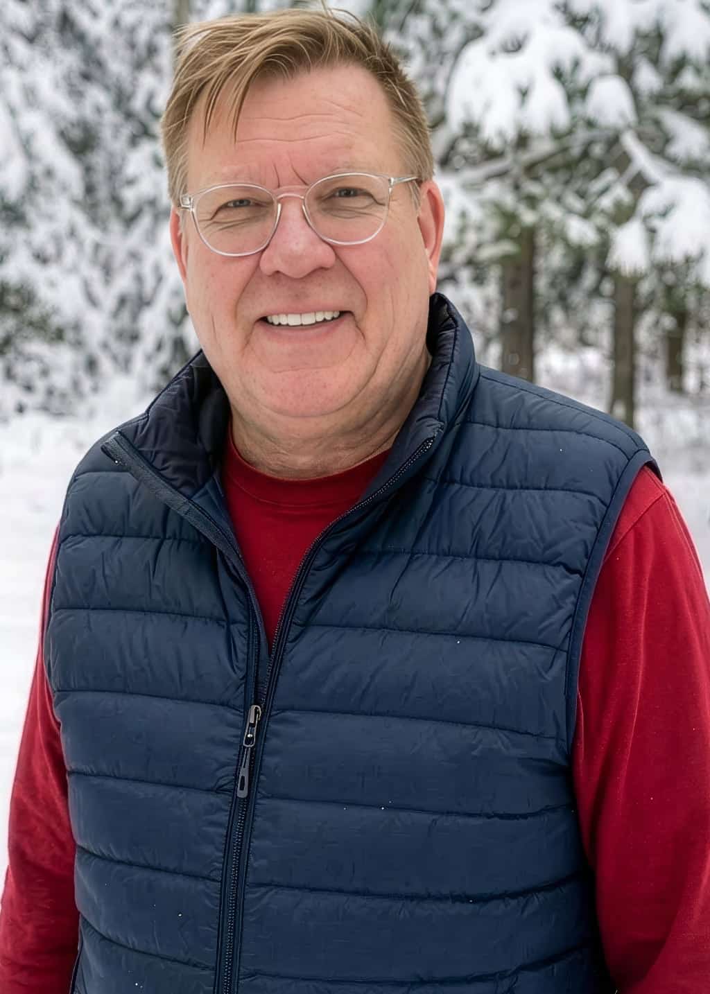 A man with light brown hair and glasses, wearing a navy blue vest over a red shirt, stands outside in a snowy forest with snow-covered trees in the background, smiling at the camera.