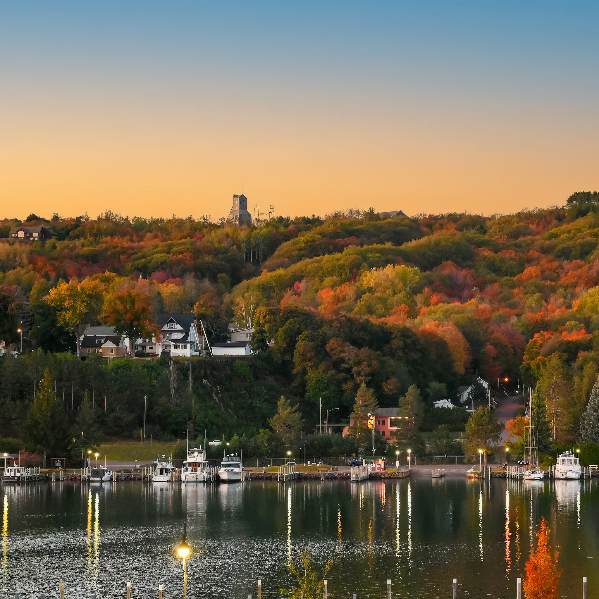 A serene lakeshore with boats docked at a marina, backed by colorful autumn trees and houses nestled on a hillside under a clear sunset sky.