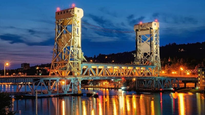 A brightly lit lift bridge spans a river at dusk, with reflections of lights shimmering on the water. The sky is partly cloudy, and the surrounding area includes trees and a few buildings.