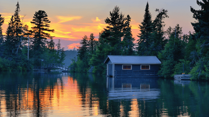 A wooden boathouse sits on a calm lake surrounded by trees at sunset, with the sky glowing orange and pink and reflections shimmering on the water. A small arched bridge is visible in the background.