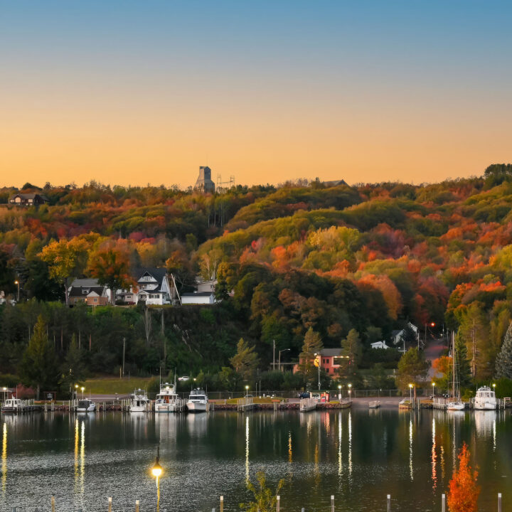 A serene lakeshore with boats docked at a marina, backed by colorful autumn trees and houses nestled on a hillside under a clear sunset sky.