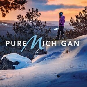 A person in a purple jacket stands on a snowy hill at sunset, overlooking a landscape of snow-covered trees and rolling hills, with Pure Michigan text across the image.