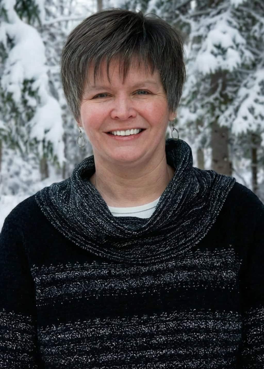 A woman with short brown hair smiles at the camera, wearing a black and gray sweater. She stands outdoors in a snowy forest with snow-covered trees in the background.