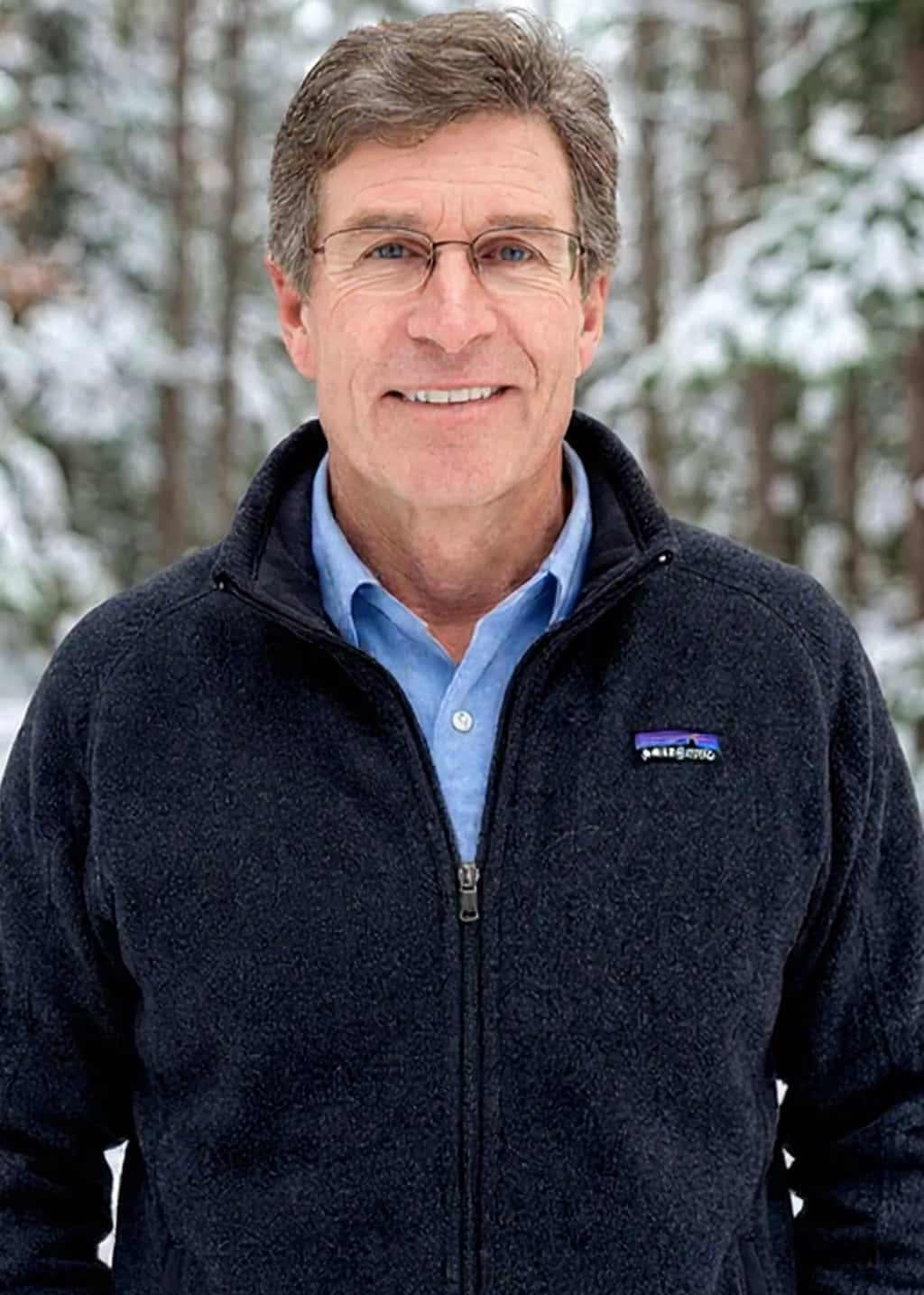 A middle-aged man with glasses and short brown hair is smiling outdoors, wearing a blue collared shirt and a dark zip-up jacket. Snow-covered trees are in the background.