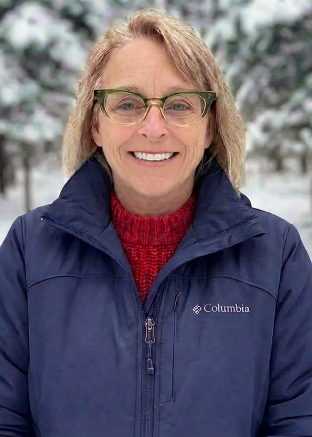 A woman with blonde hair and glasses smiles outdoors, wearing a navy Columbia jacket over a red sweater. Snow-covered trees are visible in the blurred background.