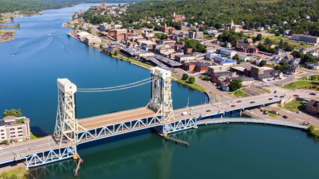 Aerial view of a lift bridge spanning a waterway, connecting two parts of a town with buildings, roads, and green hills in the background.