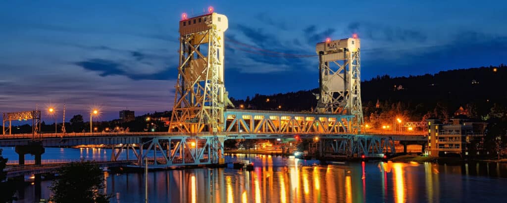 A brightly lit lift bridge spans a calm river at dusk, with its lights reflecting on the water and a twilight sky in the background. City lights and buildings are visible on both sides of the bridge.