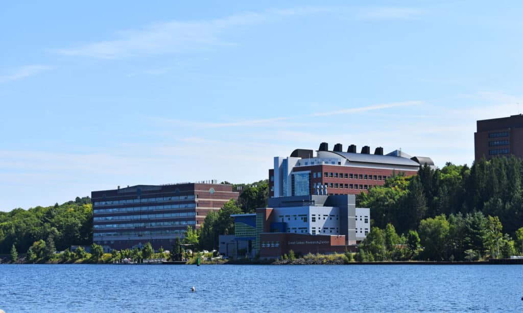 A large research center with modern buildings sits along a tree-lined riverside under a clear blue sky.