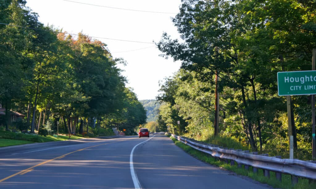 A car drives on a tree-lined road with a green sign on the right reading Houghton City Limits under a clear sky.