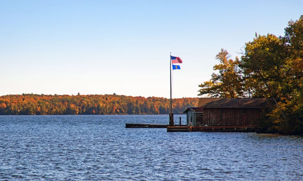 A wooden dock with American and blue flags stands by a lake. Trees with autumn foliage line the waters edge under a clear sky.