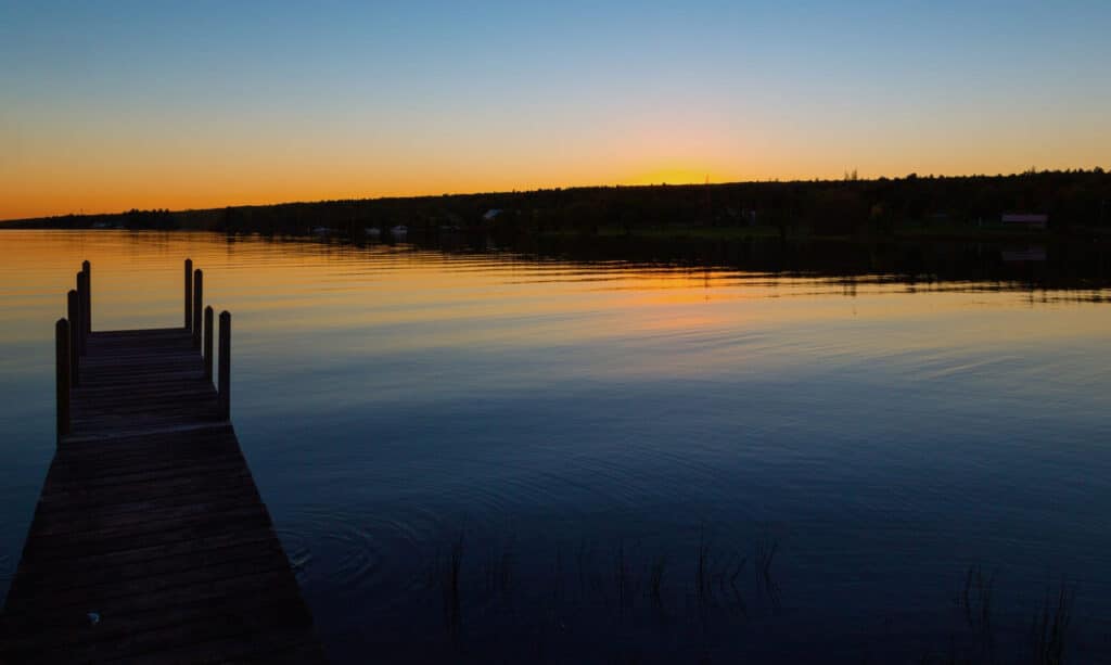 A wooden dock extends into a calm lake at sunset, with orange and blue hues reflected on the water and a tree-lined shore in the distance.