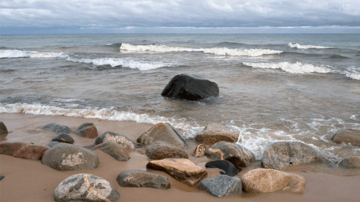 Rocky shoreline with various large stones on the sandy beach, gentle waves rolling in from a cloudy, distant horizon across the sea.