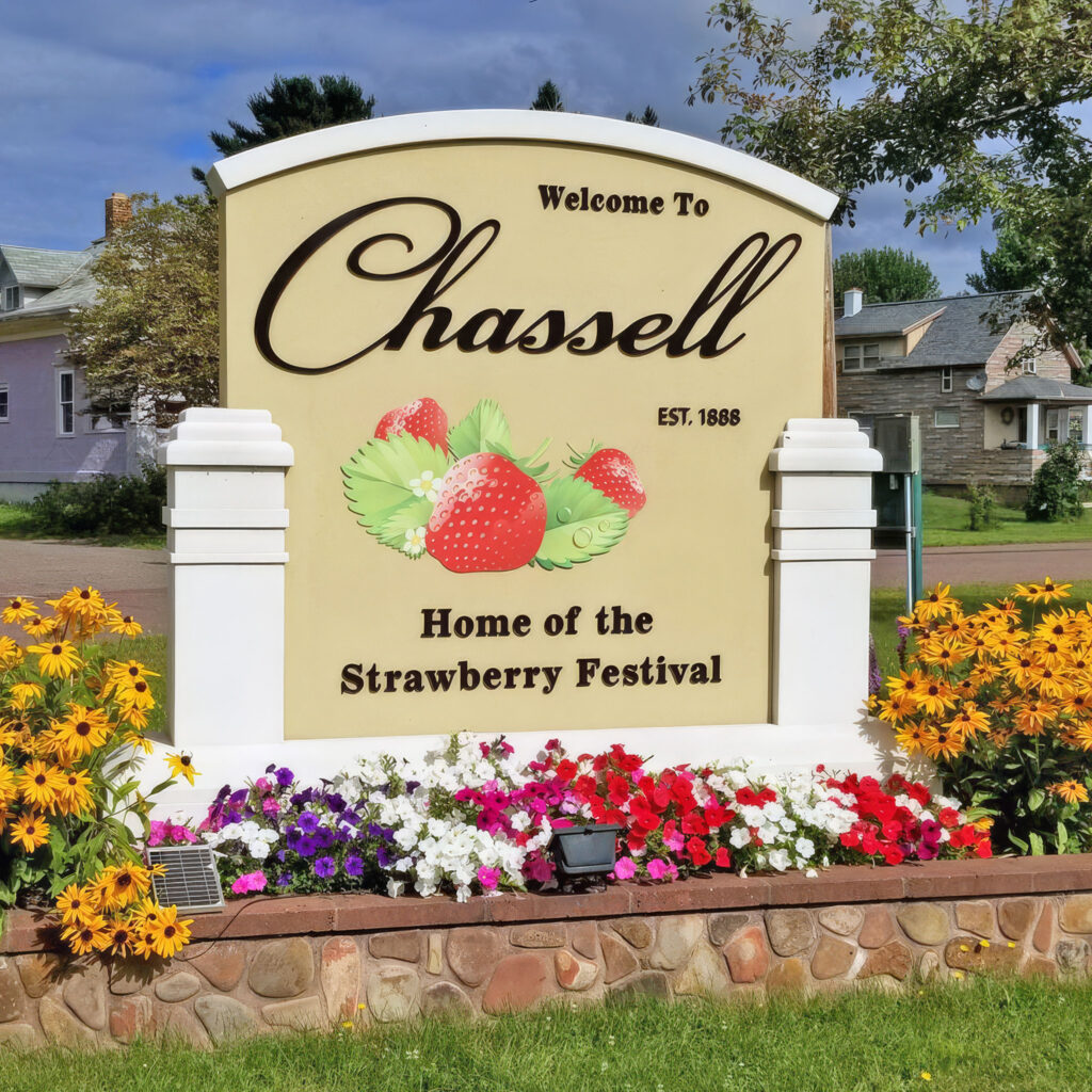 A large welcome sign for Chassell MI, decorated with strawberries and surrounded by colorful flowers. The sign reads, Welcome to Chassell, EST. 1888, Home of the Strawberry Festival.