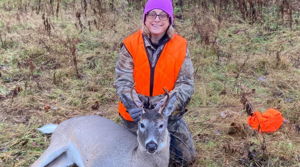 Kathy-Liimatta-Hunting-Deer- | State Wide Real Estate of Houghton A person wearing glasses, a purple hat, and an orange hunting vest kneels in tall grass, smiling and holding the antlers of a large deer lying on the ground.