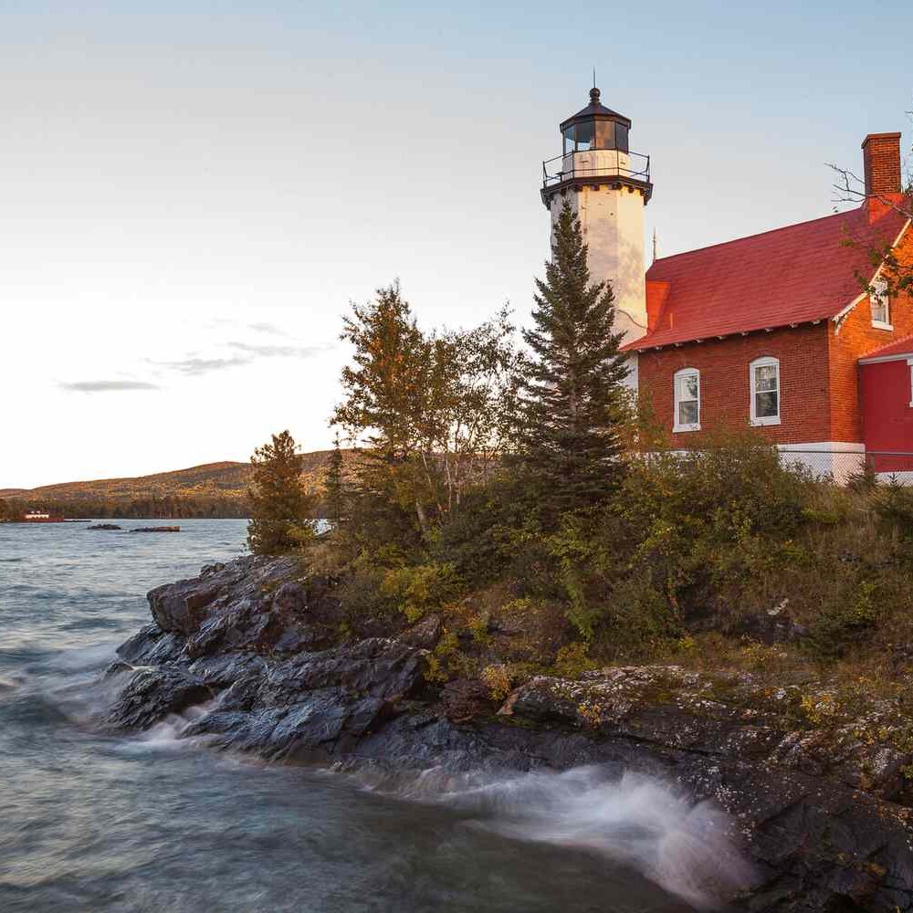 A red-roofed lighthouse and brick keeper’s house sit on a rocky shoreline beside a lake, surrounded by trees, with calm water and hills in the background under a clear sky at sunset.
