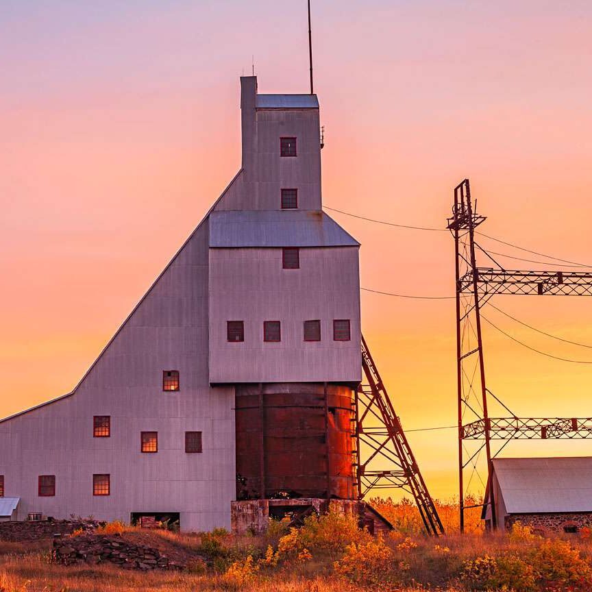 A tall, angular mining building with a metal exterior stands against a colorful sunset sky, with two large metal towers and another small structure nearby in a field.