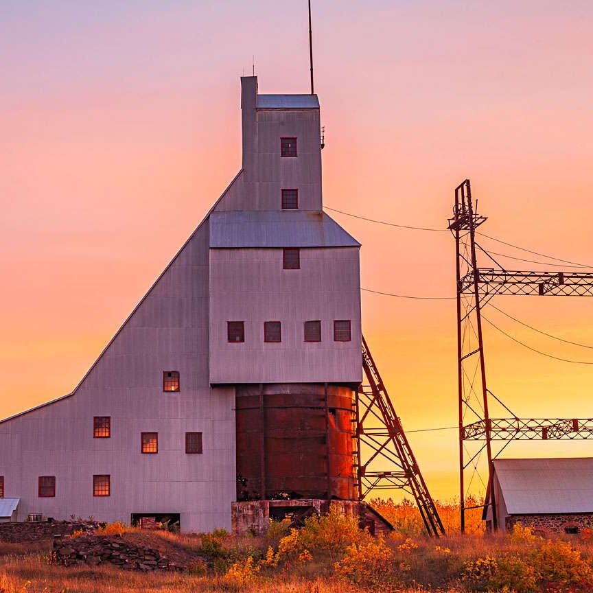A tall, angular mining building with a metal exterior stands against a colorful sunset sky, with two large metal towers and another small structure nearby in a field.