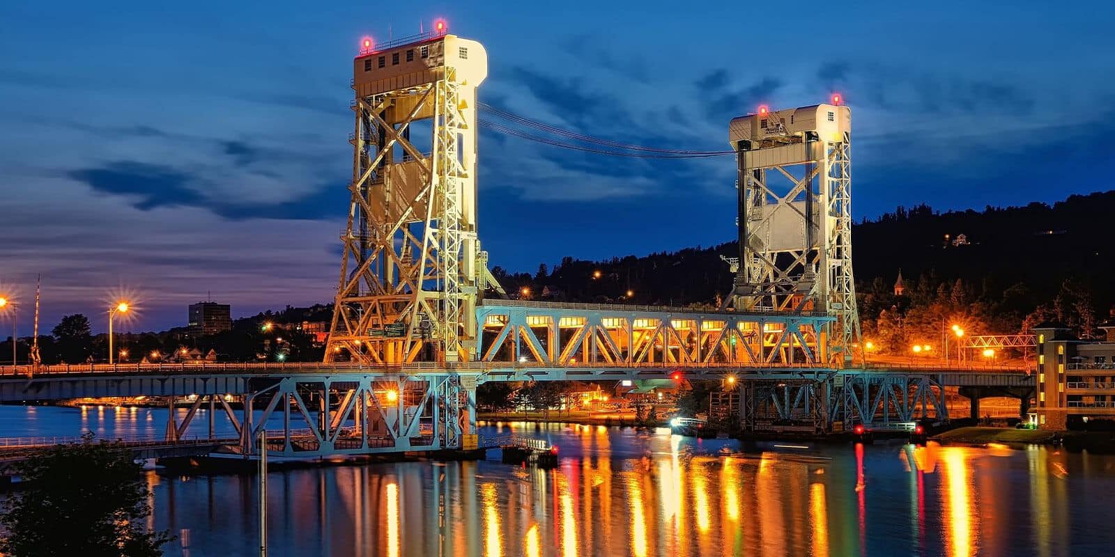 A brightly lit lift bridge spans a calm river at dusk, with its lights reflecting on the water and a twilight sky in the background. City lights and buildings are visible on both sides of the bridge.