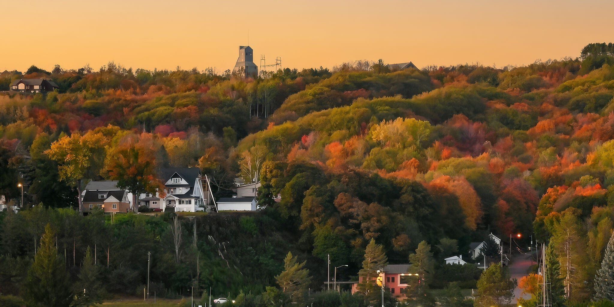 A serene lakeshore with boats docked at a marina, backed by colorful autumn trees and houses nestled on a hillside under a clear sunset sky.