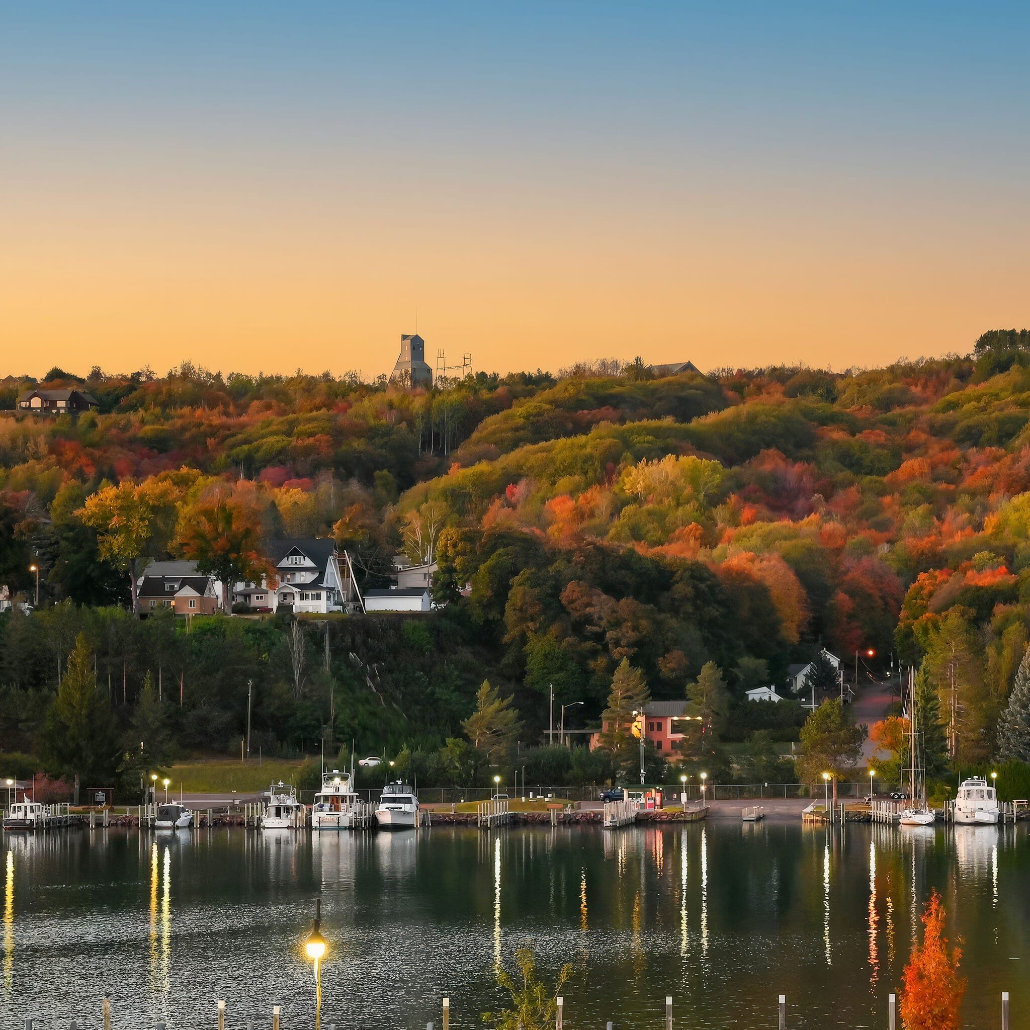 A serene lakeshore with boats docked at a marina, backed by colorful autumn trees and houses nestled on a hillside under a clear sunset sky.
