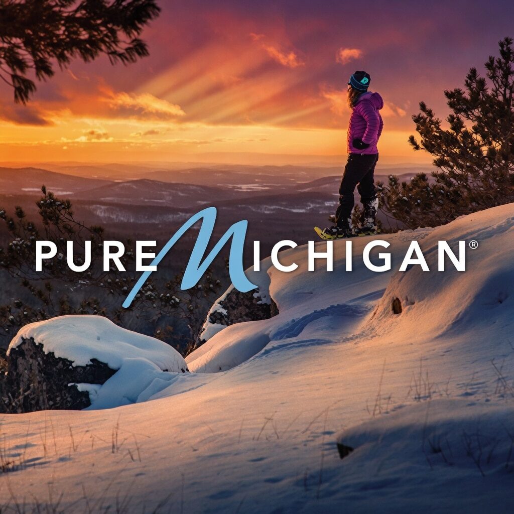 A person in winter gear stands on a snowy hill at sunset, overlooking a scenic landscape of snow-covered trees and distant hills. The words Pure Michigan are boldly displayed across the image.