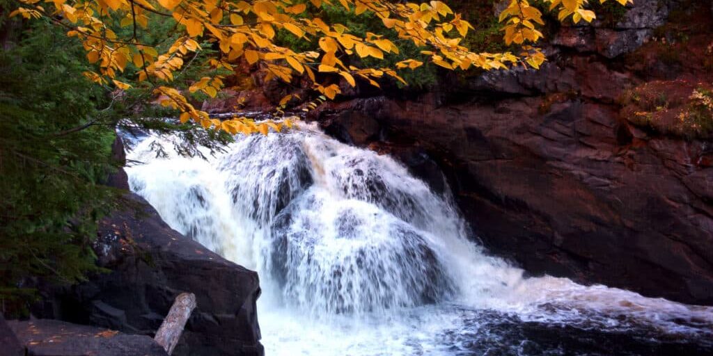 Waterfall cascades over dark rocks surrounded by green foliage and yellow autumn leaves, creating a vibrant natural scene.