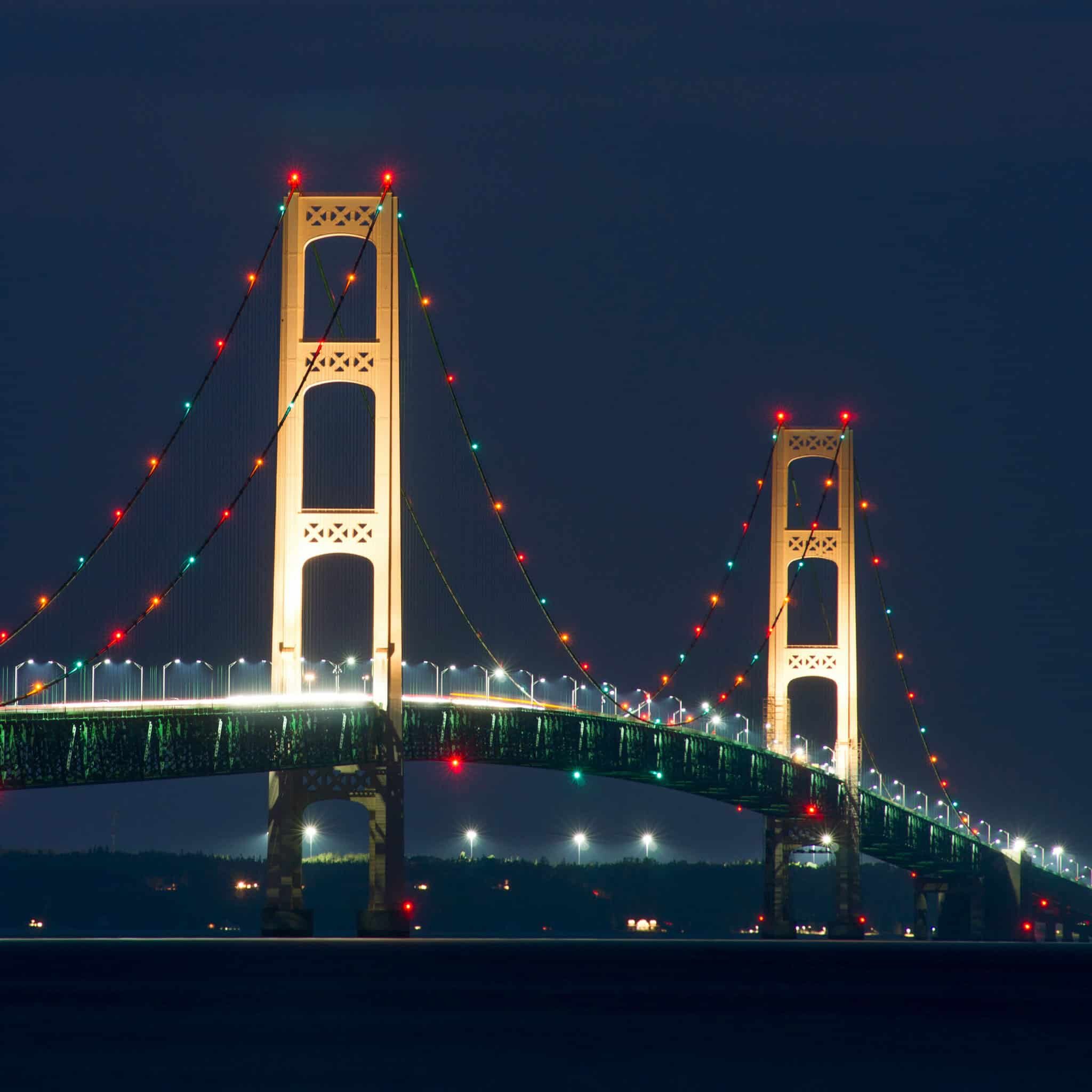 A large suspension bridge illuminated at night with colorful lights along its cables and towers, spanning across a body of water against a dark sky.