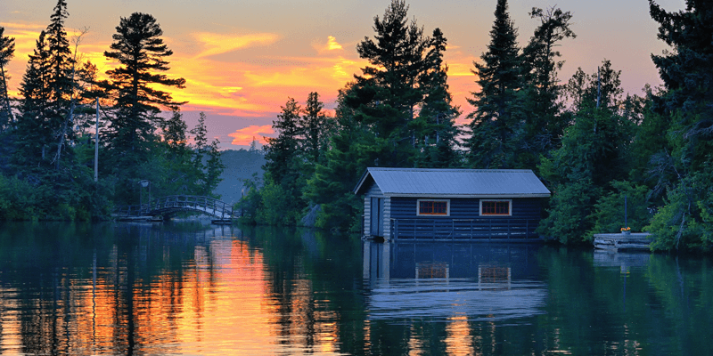 A wooden boathouse sits on a calm lake surrounded by trees at sunset, with the sky glowing orange and pink and reflections shimmering on the water. A small arched bridge is visible in the background.