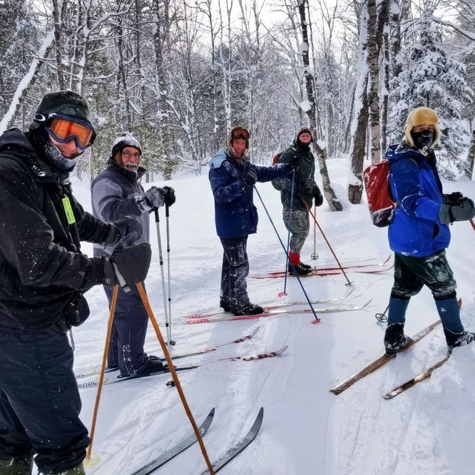 Five people in winter clothing stand on skis in a snowy forest, holding ski poles. Snow covers the ground and trees, and the group appears to be taking a break or posing for a photo on a skiing trip.